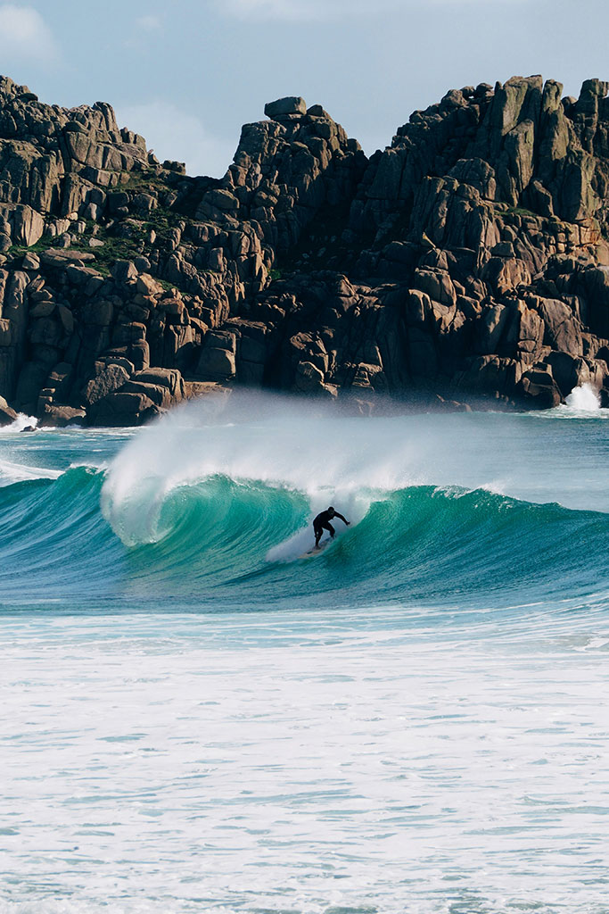 Expert surfer drawing a line beneath a pitching Cornish wave backed by dark granite cliffs