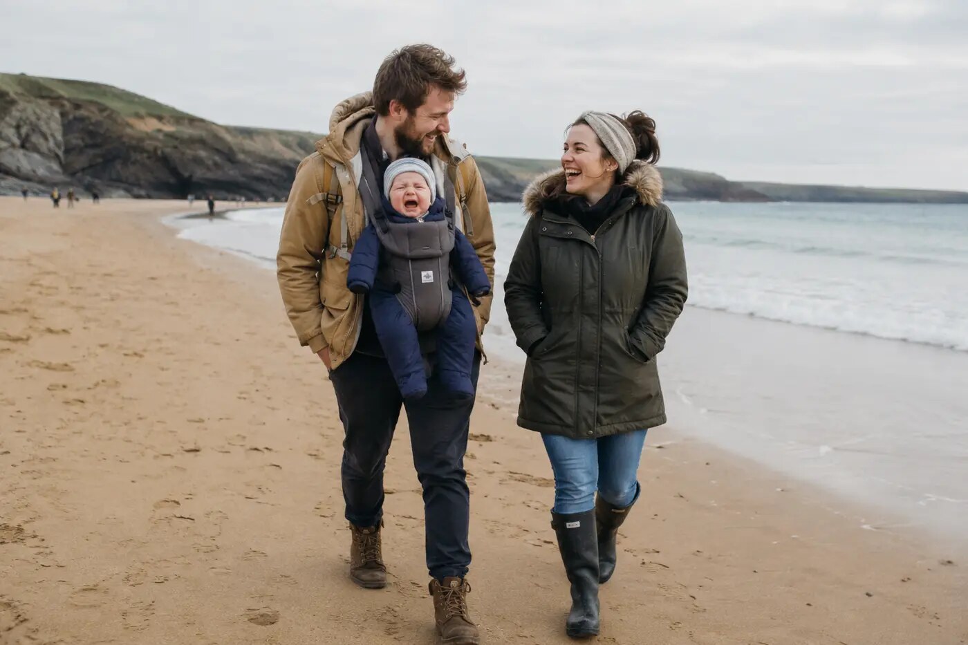 Parents laughing together while carrying their crying baby along a Cornish beach