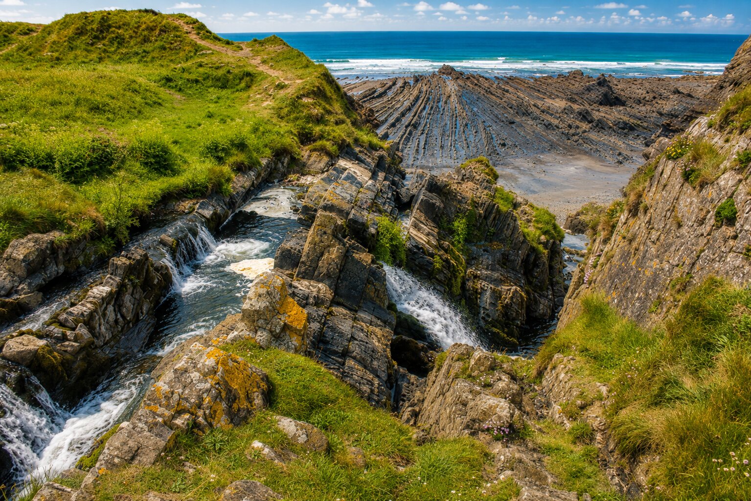 Welcombe Mouth waterfall cascading through folded rocks towards the blue Atlantic in summer sunshine