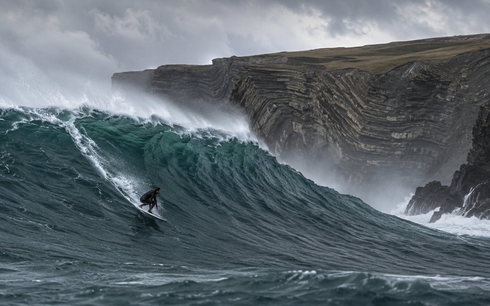 Representative cold-water surfer riding a powerful dark Atlantic wave below folded coastal cliffs